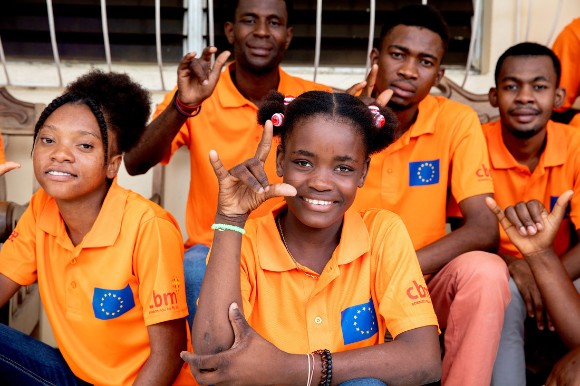 Five teenagers sit in a group looking at the camera, making positive hand gestures. They wear orange t-shirts with the CBM and EU logos.